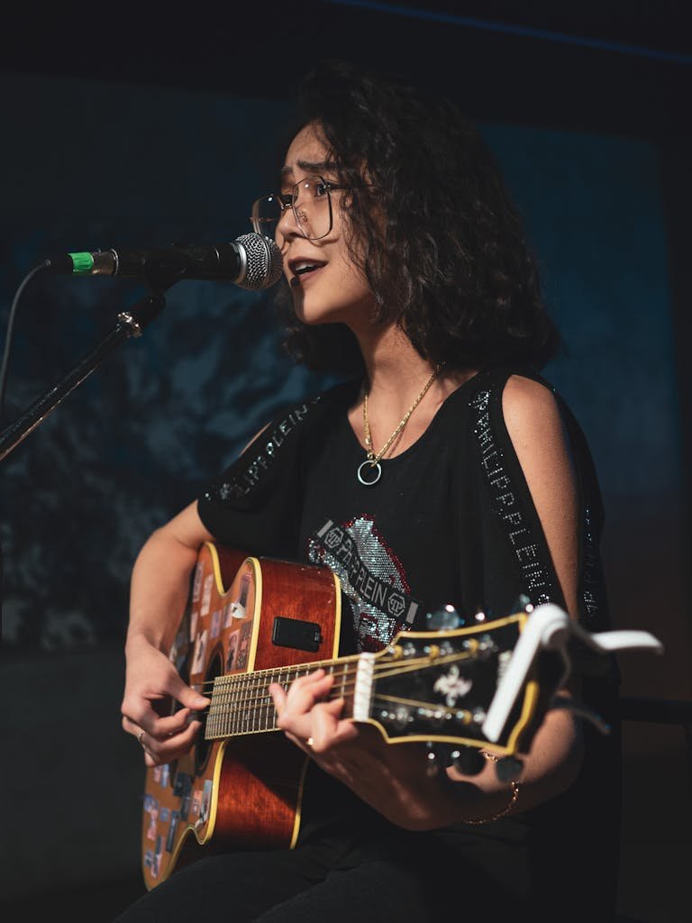 Young woman with eyeglasses singing and playing acoustic guitar on stage.